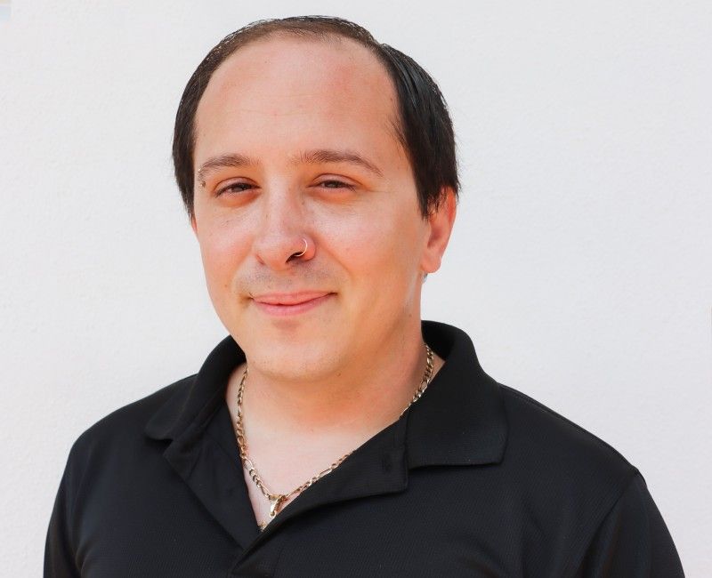 A person with a nose piercing and a short hairstyle, wearing a black collared shirt, smiles slightly.